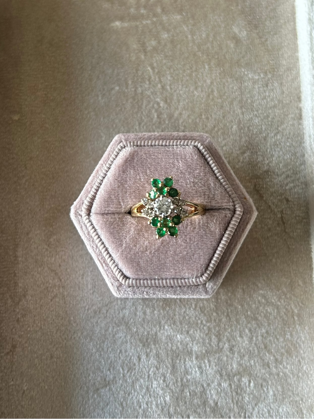 A yellow gold ring with a green and white floral cluster center, displayed on a hexagonal pink velvet ring box inside an octagonal cream-colored presentation box.