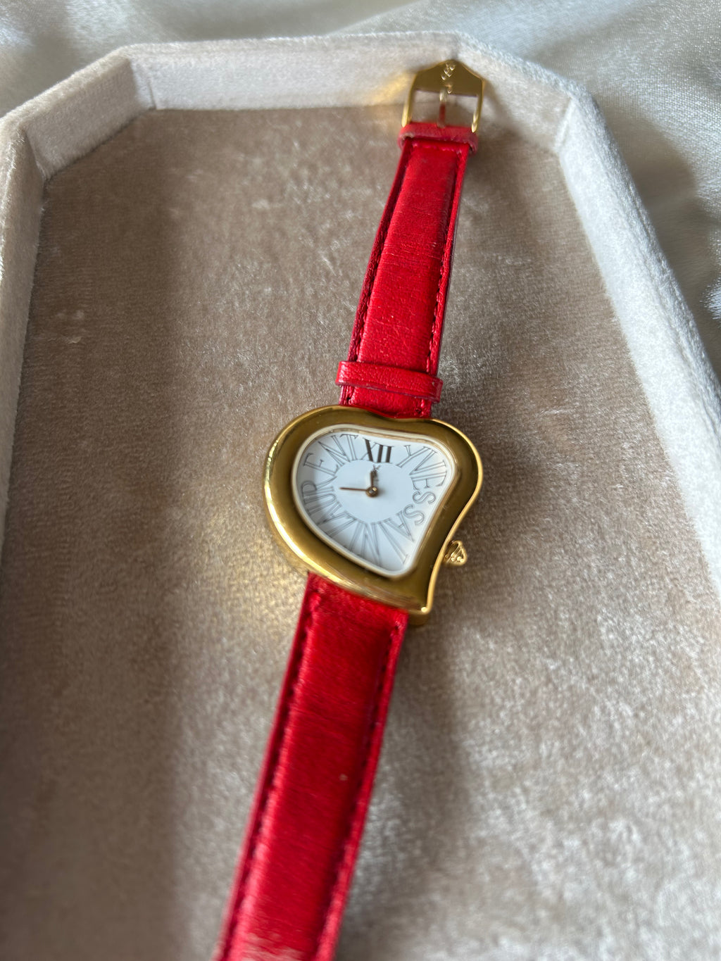 A vintage-style wristwatch with a gold-tone heart-shaped case, white dial, and bright red strap, resting inside a beige presentation box.