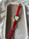 A vintage-style heart-shaped watch with a bright red leather strap and gold-tone case, displayed inside an open cream-colored presentation box on a soft beige fabric background.