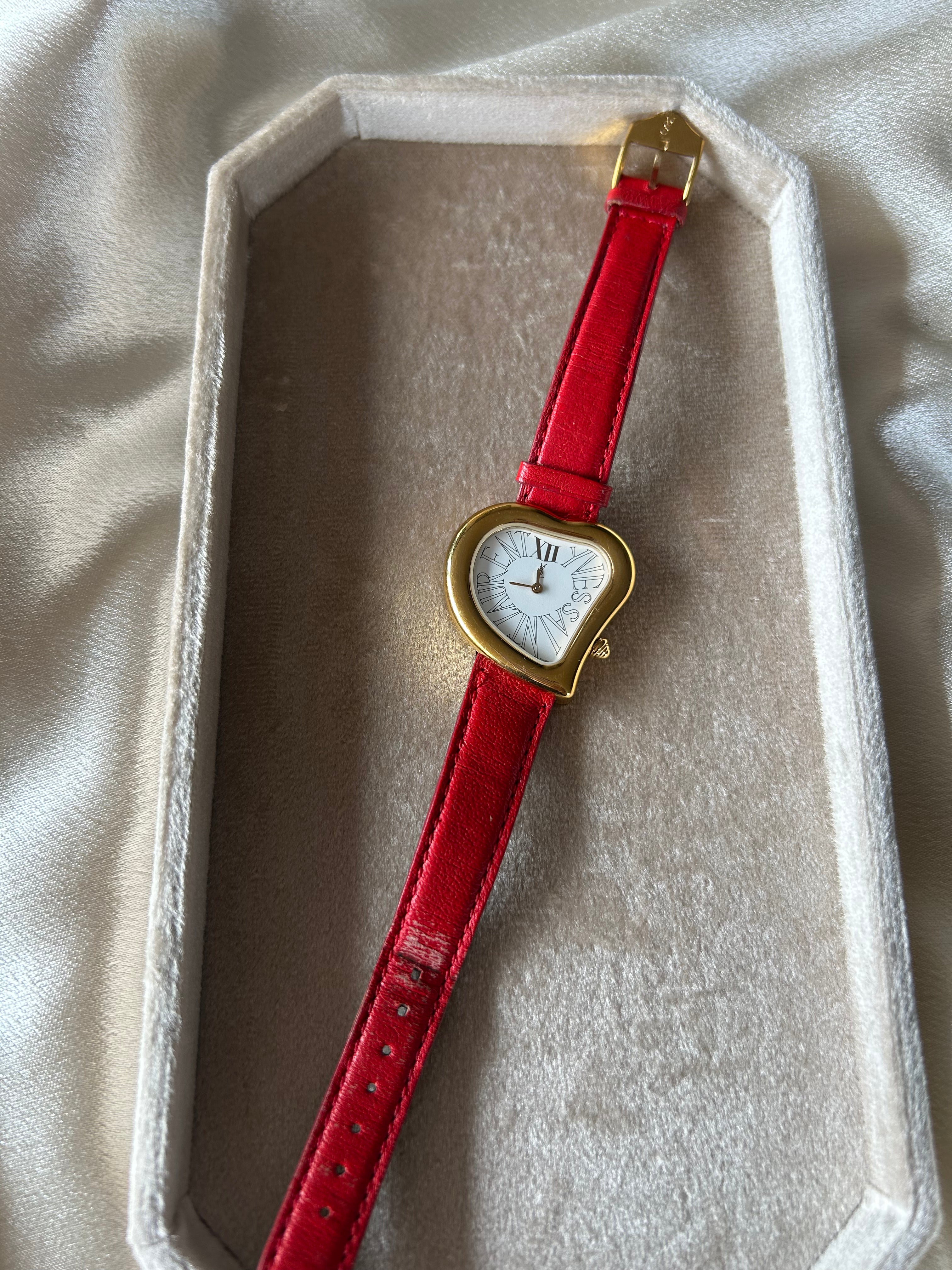 A vintage-style heart-shaped watch with a bright red leather strap and gold-tone case, displayed inside an open cream-colored presentation box on a soft beige fabric background.